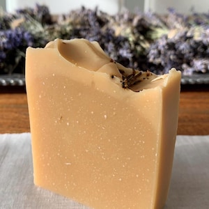 May include: A light tan, rectangular bar of handmade soap with flecks. The top surface is uneven and textured, with small, dark inclusions. Dried lavender is visible in the background, suggesting a lavender-scented soap. The soap is likely scented.