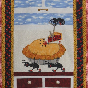 May include: A quilted wall hanging with a cherry pie theme. Three black ants are carrying a large cherry pie with a slice missing. The pie is on a white surface with a window behind it. The wall hanging has a red and white checkered border.