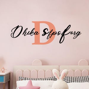 Könnte beinhalten: Wandaufkleber mit dem Text "Ohika Sffpsforg" in schwarzer Schrift und einem großen korallenfarbenen Buchstaben "D". Der Aufkleber befindet sich an einer rosa Wand über einem Bett mit dekorativem Kopfteil und Kissen.