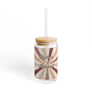 May include: A clear glass tumbler with a bamboo lid and a clear straw. The tumbler features a retro design with the words "Hooked & Barred" in a vintage font. The design includes a sunburst pattern in shades of brown, beige, and red.