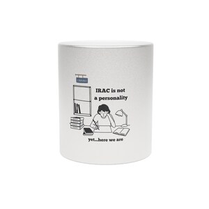 May include: A silver-coloured coffee mug with a black and white illustration. The illustration shows a person at a desk with books, a lamp, and the text "IRAC is not a personality, yet...here we are."