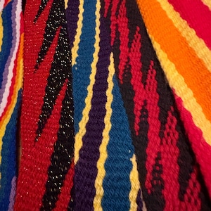 May include: A collection of woven fabric strips featuring vibrant, multi-colored patterns. The designs include vertical stripes in red, orange, yellow, blue, and purple, alongside zigzag motifs in black and red with subtle metallic thread accents.