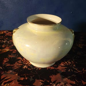 Op de afbeelding: Crèmekleurige keramische vaas met een afgeronde body en een brede opening. De vaas heeft gouden accenten op de rand en handvatten. Hij staat op een patroon oppervlak tegen een donkerblauwe achtergrond.