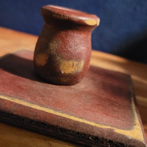 May include: A small, brown wooden stamp with a rounded top, resting on a rectangular, reddish-brown wooden base. The stamp has a worn appearance, with visible wood grain and lighter patches. The base has a darker border.