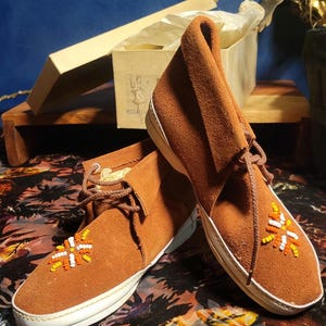 May include: Brown suede moccasin boots with white soles and beaded detailing. The boots feature a lace-up design and a folded-over top. A box with tissue paper is visible in the background.
