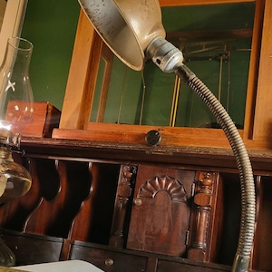 May include: An antique desk lamp with a flexible, ribbed neck and a weathered, metallic shade. The lamp is positioned in front of a dark wooden cabinet with decorative carvings. A vintage book is visible.
