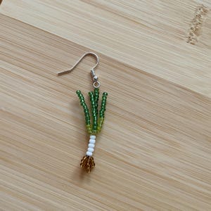 May include: A single handmade earring featuring a beaded design resembling a green onion. The earring has a silver hook and is crafted with green, yellow, white, and gold beads. The earring is set against a light brown wooden surface.