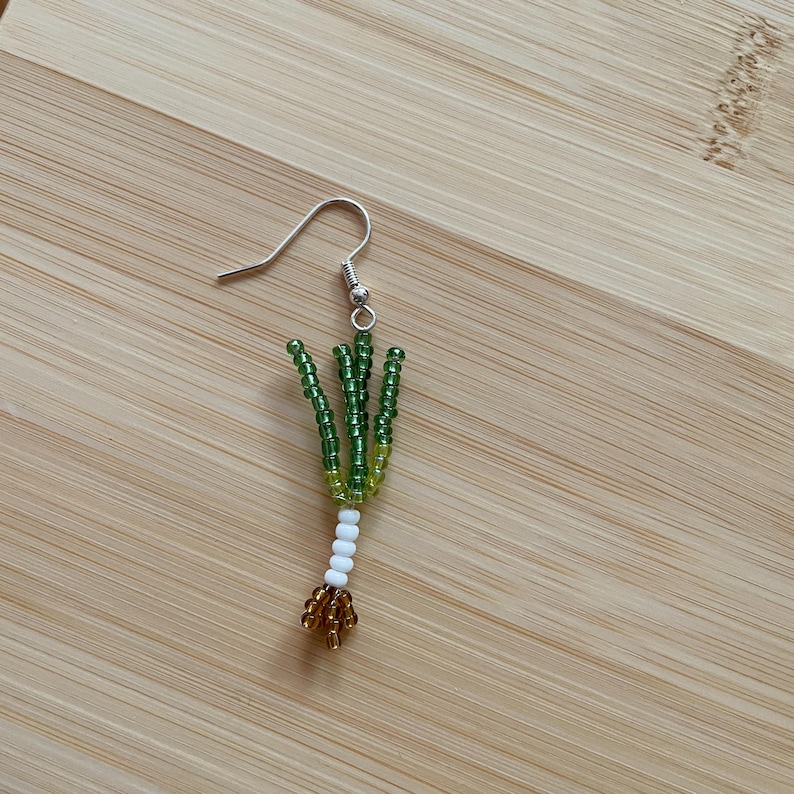 May include: A single handmade earring featuring a beaded design resembling a green onion. The earring has a silver hook and is crafted with green, yellow, white, and gold beads. The earring is set against a light brown wooden surface.