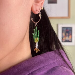 May include: A silver hoop earring with a beaded cactus design. The cactus features green beads for the body, white beads for the stem, and gold beads at the base. The earring is worn on the ear.