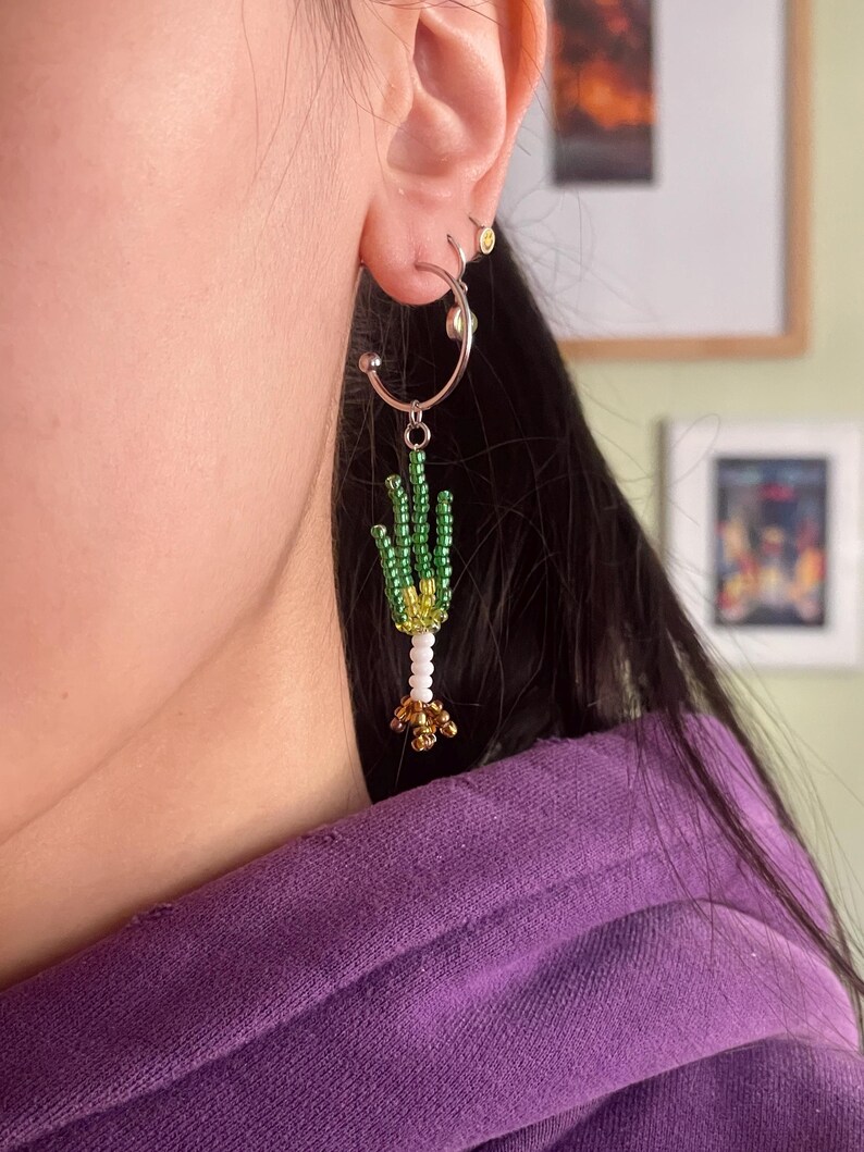 May include: A silver hoop earring with a beaded cactus design. The cactus features green beads for the body, white beads for the stem, and gold beads at the base. The earring is worn on the ear.