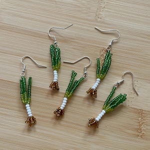 May include: Five pairs of beaded earrings shaped like green onions. Each earring features green beads for the leaves, white beads for the stalk, and brown beads for the roots. The earrings have silver-colored hooks.