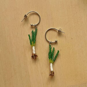 May include: A pair of silver hoop earrings, each adorned with a beaded leek design. The leeks feature green, white, and gold beads, creating a unique and whimsical accessory. The earrings are set against a light wooden background.