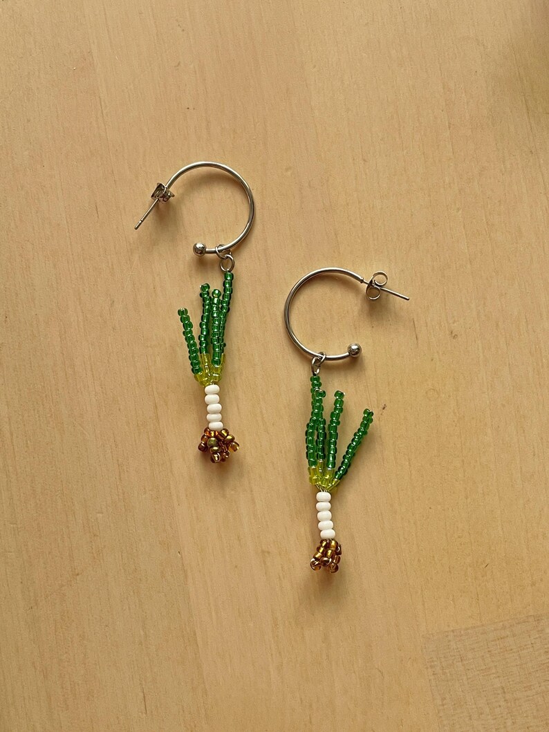 May include: A pair of silver hoop earrings, each adorned with a beaded leek design. The leeks feature green, white, and gold beads, creating a unique and whimsical accessory. The earrings are set against a light wooden background.