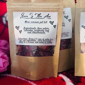 Puede incluir: Bolsas de papel kraft etiquetadas "Love Is The Air" que contienen mini kits de olla a fuego lento. Los kits incluyen pétalos de rosa, lavanda, hojas de frambuesa y fresas. Las instrucciones están impresas en la etiqueta. El fondo es rojo.