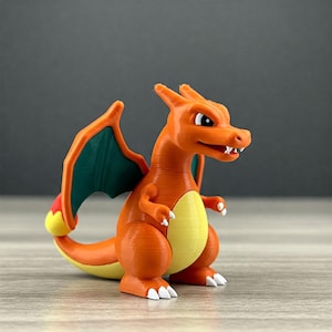 Könnte beinhalten: Eine 3D-gedruckte orangefarbene Charizard-Figur mit grünen Flügeln und einem gelben Bauch. Der Drache hat weiße Krallen und Zähne und steht auf einer Holzoberfläche. Die Figur ist ein Sammlerobjekt.