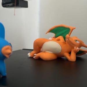 May include: An orange Charizard figurine with green wings and a white belly is lying down, with a blue and orange figurine in the foreground. The Charizard is in a relaxed pose, resting its head on its hand.