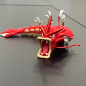 May include: A red and white figurine of a serpentine creature with an open mouth, displaying sharp teeth and a pink tongue. The figurine has a long body with segmented sections and white fins. The creature's head features prominent red spikes and a white crest.