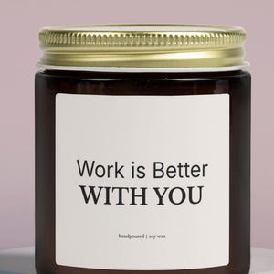 Puede incluir: Una vela en tarro de cristal marrón con tapa dorada, con una etiqueta blanca con el texto "Work is Better WITH YOU". La vela está sobre una plataforma blanca en capas sobre un fondo rosa suave.