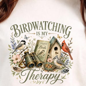 Op de afbeelding: Crèmekleurig T-shirt met de tekst "Birdwatching is my Therapy" in groene letters. De afbeelding toont een vogelhuisje, een verrekijker, een veldgids en diverse vogels en bloemen, wat een ontwerp met een natuurthema creëert.