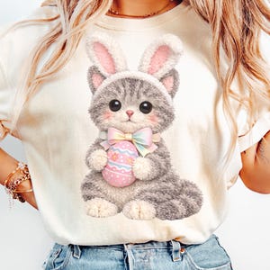 Puede incluir: Camiseta color crema con un lindo gato de dibujos animados disfrazado de conejito, sosteniendo un huevo de Pascua decorado. El gato tiene pelaje gris y blanco, ojos grandes y un lazo rosa. Las orejas de conejo son rosas y grises. La camiseta se combina con unos vaqueros azules.