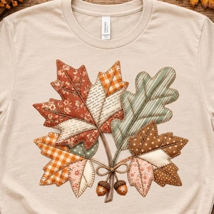 Puede incluir: Una camiseta beige de manga corta con un gráfico central de un grupo de hojas de otoño. Las hojas están representadas en un estilo de patchwork con varios patrones que incluyen flores, cuadros, lunares y texto, atadas con dos pequeñas bellotas.