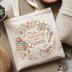 May include: Cream-colored tote bag featuring a floral wreath design with the words "Quilting Queen." The design includes images of sewing tools, fabric, and spools of thread. The bag has two beige handles.