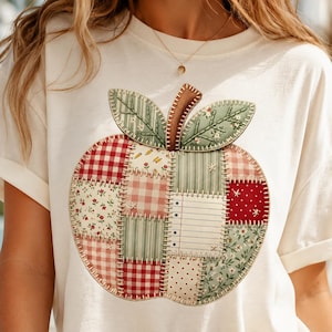 May include: A cream-colored t-shirt featuring a large, central apple graphic composed of various patchwork-style fabric squares. The apple design includes patterns like red gingham, floral prints, and lined paper, all outlined with visible white stitching.