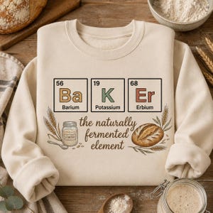 May include: A cream-colored crewneck sweatshirt featuring a graphic design with three periodic table squares spelling "BaK Er" for Barium, Potassium, and Erbium. Below, it reads "the naturally fermented element" with a sourdough starter jar and bread.