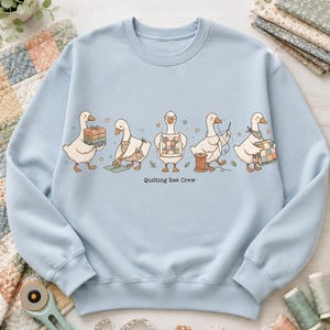 May include: A light blue crewneck sweatshirt featuring a graphic of five white geese engaged in quilting activities, with the text "Quilting Bee Crew" printed below. The geese are shown holding fabric stacks, using a rotary cutter, displaying a quilt block, sewing with a needle and thread, and carrying a finished quilt.