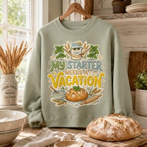 May include: A sage green crewneck sweatshirt featuring a graphic print of a sourdough starter jar wearing sunglasses in a hammock, a loaf of bread, a whisk, and a rolling pin, with the text "MY STARTER NEEDS A VACATION" in colorful lettering.