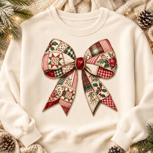 May include: A cream-colored crewneck sweatshirt featuring a large, central graphic of a decorative bow. The bow is composed of various patchwork patterns including red and green plaid, holly berries, floral motifs, and star-shaped quilt designs.