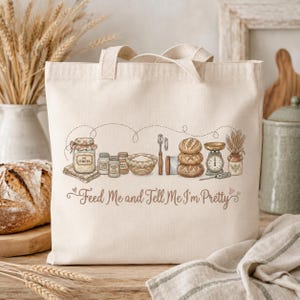 Może przedstawiać: Kremowa torba typu tote z ilustracją o tematyce piekarniczej. Projekt zawiera słoiki, miski, chleb, wagę i napis "Feed Me and Tell Me I'm Pretty". Torba ma dwa uchwyty.