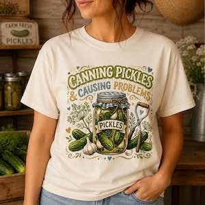 May include: Cream-colored t-shirt featuring a graphic of a pickle jar with the text "CANNING PICKLES & CAUSING PROBLEMS." The design includes pickles, garlic, and dill. A vintage-style sign that says "FARM FRESH PICKLES" is in the background.