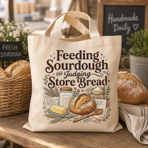 Puede incluir: Una bolsa de tela beige con un estampado gráfico en marrón oscuro que dice Feeding Sourdough and Judging Store Bread. La ilustración incluye una hogaza de pan, un frasco de masa madre, un trozo de mantequilla, harina y tarjetas de recetas.