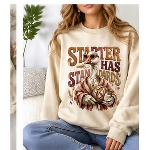 May include: A cream-colored crewneck sweatshirt featuring a graphic of a goose wearing sunglasses and a necklace, holding a loaf of bread. The text reads STARTER HAS STANDARDS in brown and red lettering, with wheat stalks surrounding the goose.