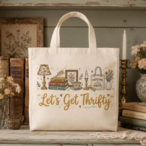 May include: Cream-colored tote bag with a whimsical illustration of vintage items, including a lamp, books, and a teacup. The phrase "Let's Get Thrifty" is written in gold script. Perfect for carrying books, groceries, or other items.