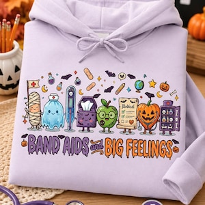 May include: A light purple hooded sweatshirt featuring a colorful graphic print of various medical-themed characters and items, including a mummy, ghost, thermometer, and pumpkin. The text "BAND AIDS and BIG FEELINGS" is printed in purple and orange.