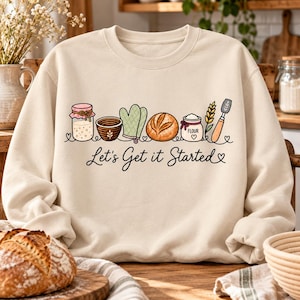 May include: A cream-colored crewneck sweatshirt featuring a graphic illustration of baking supplies including a jar, bowl, oven mitt, loaf of bread, flour container, wheat stalk, and spatula, with the text Let's Get it Started written below.