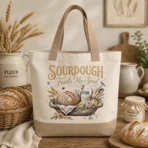 Może przedstawiać: Kremowa torba płócienna z beżowymi uchwytami i podstawą. Torba zawiera napis "SOURDOUGH Feeds My Soul" z akwarelową ilustracją bochenka chleba, kwiatów i przyborów do pieczenia. Uroczy dodatek.