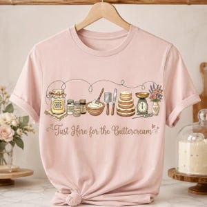 May include: Pale pink t-shirt with a baking-themed graphic. The design includes a jar of butter, a whisk, and a cake, with the text "Just Here for the Buttercream". The shirt is displayed on a wooden hanger, ready to wear.