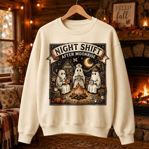 May include: A cream-colored crewneck sweatshirt featuring a graphic print of three ghosts sitting around a campfire. The text reads "NIGHT SHIFT AFTER MOONRISE" above a scene with a hospital building, pumpkins, and mugs. The design uses warm earth tones.
