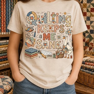 May include: Beige t-shirt with the text "Quilting Keeps Me Sane" in a patchwork design. The graphic includes quilting tools, a goose, and a basket of fabric, perfect for quilters and sewing enthusiasts.