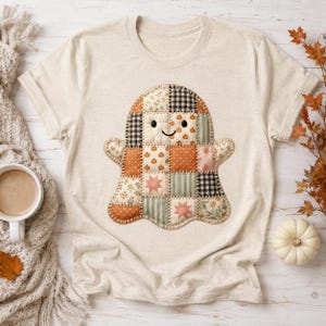 May include: A cream-colored t-shirt featuring a central graphic of a ghost constructed from a patchwork quilt design. The ghost is composed of various fabric squares in shades of orange, green, and black, displaying patterns of pumpkins, stars, and floral motifs with black stitched outlines.