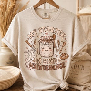 Puede incluir: Camiseta beige con un gráfico que dice "MY STARTER IS HIGH MAINTENANCE". El diseño presenta un tarro sonriente de masa madre, espigas de trigo y una cuchara de madera. También se incluye una pequeña "Sourdough Guide".