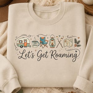 May include: A beige crewneck sweatshirt featuring a graphic print of camping items including a camper, a coffee mug labeled Camp Coffee, a folding chair, a lantern, marshmallows on sticks, a map, and pine branches with the text Let's Get Roaming.