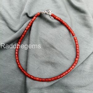 May include: A red beaded necklace with a silver clasp. The necklace is made of small, square-shaped beads. The background is a gray fabric. The text "Raddhegems" is visible in the lower left corner.
