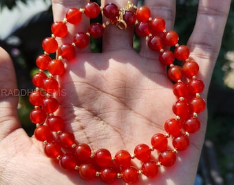 May include: A double-strand necklace featuring vibrant red carnelian beads. The beads are strung with gold-colored spacers and clasps. The necklace is displayed on a hand, showcasing the rich color and design of the jewelry.