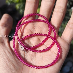 May include: A delicate necklace composed of small, faceted, magenta-colored beads. The necklace is strung together with a silver clasp. The image is taken in natural light, highlighting the vibrant color of the beads. The text "Raddhegems" is visible in the lower left corner.