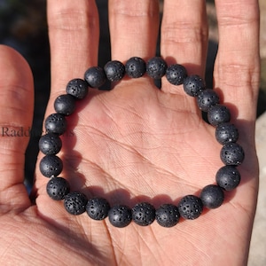 Può includere: Braccialetto con perline di roccia lavica nera. Il braccialetto presenta perline rotonde e porose infilate su un elastico. Le perline sono di colore antracite scuro e il braccialetto è mostrato su una mano.