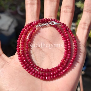 Puede incluir: Un delicado collar compuesto por pequeñas cuentas facetadas de color rojo intenso. El collar está unido con un cierre plateado. El texto "Raddhegems" es visible. Este collar es una joya.